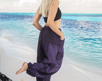 May include: A woman wearing a black tank top and purple printed pants with a geometric pattern. She is standing on a white sandy beach with blue water in the background.
