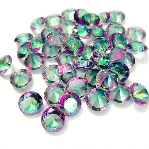 May include: A collection of iridescent, round-cut gemstones. The gems display a spectrum of colors, including green, purple, and blue, with a faceted surface that catches the light. The gemstones are scattered on a white background.