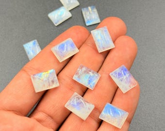 May include: A collection of rectangular moonstone cabochons. The stones display a milky white base with iridescent blue and rainbow flashes. The gemstones are held in a hand against a gray background. These stones are suitable for jewelry making.