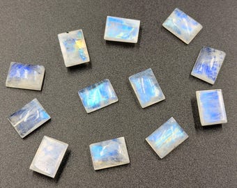 May include: A collection of rectangular moonstone cabochons. Each stone displays a milky white base with iridescent blue and yellow flashes. The stones are arranged on a dark gray surface, showcasing their polished appearance.