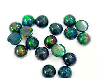 May include: A selection of polished, round black opal gemstones displaying flashes of green, red, and orange. The opals are scattered on a white surface, highlighting their iridescent colours. These gemstones are suitable for jewellery making.