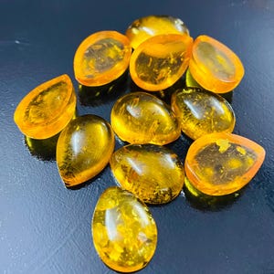 May include: A selection of polished amber gemstones in diverse shapes, including teardrops and ovals. The stones are a translucent golden-yellow hue, with visible internal patterns. The gemstones are arranged on a dark, reflective surface.