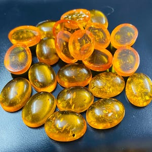 May include: A selection of polished amber cabochons in various oval and rounded shapes. The translucent stones display warm orange and golden hues, with visible internal textures. The cabochons are scattered across a dark surface.