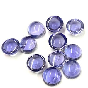 Peut inclure: Dix pierres précieuses rondes en verre violet translucide sont disposées sur une surface blanche. Chaque gemme a un dessus bombé et un fond plat, reflétant la lumière. Les gemmes sont de couleur violet profond.
