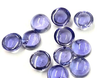 May include: A collection of ten translucent, round, purple glass gems. Each gem has a domed top and a flat bottom, reflecting light. The gems are scattered on a white surface, creating a visually appealing arrangement.