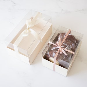 May include: Two clear plastic gift boxes with lids. The larger box is empty and tied with a cream-colored ribbon. The smaller box contains four chocolate-covered cookies tied with a pink ribbon.