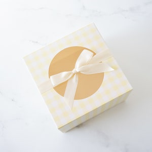 May include: A yellow and white checkered gift box with a circular window and a white satin ribbon tied in a bow.