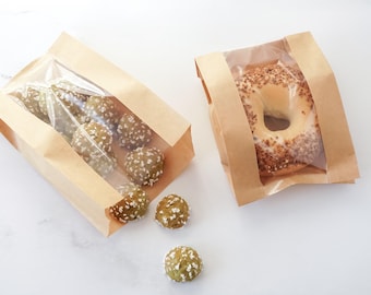May include: Two brown paper bags with clear windows. One bag contains several green, round treats covered in white sprinkles. The other bag holds a bagel covered in seeds. The bags are on a white surface.