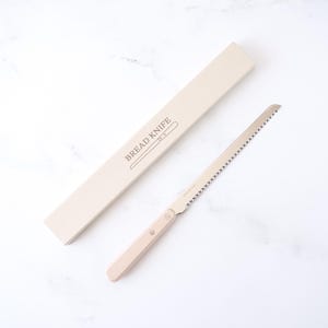 May include: A bread knife with a light wood handle and a serrated stainless steel blade. The knife is next to a beige box with the words "BREAD KNIFE" printed on it. The knife is designed for slicing bread.