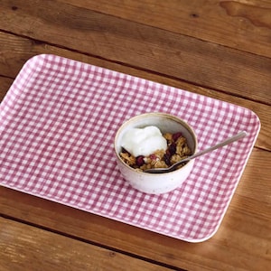 May include: A rectangular serving tray with a pink and white gingham pattern. A small ceramic bowl filled with granola and yogurt sits on the tray, along with a silver spoon. The tray rests on a wooden surface.