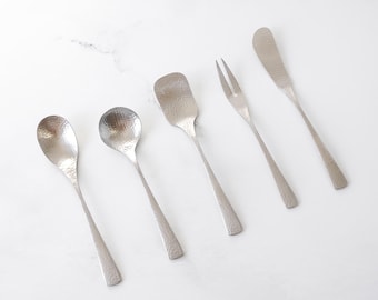 Könnte beinhalten: Ein Set aus fünf gehämmerten Servierutensilien aus Edelstahl. Das Set beinhaltet einen Löffel, einen runden Löffel, einen Löffel mit quadratischem Ende, eine Gabel und ein Buttermesser. Die Utensilien sind auf einer weißen Oberfläche angeordnet.