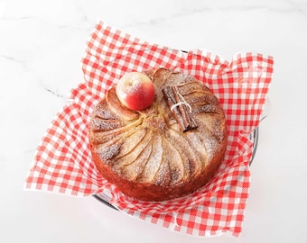 May include: A round apple cake with sliced apples arranged on top, dusted with powdered sugar. A small red apple and a cinnamon stick are placed on top. The cake sits on a red and white checkered paper.