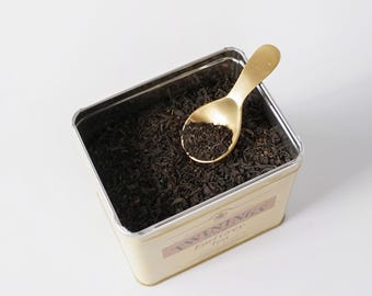 Könnte beinhalten: Eine rechteckige Metalldose gefüllt mit losem schwarzem Tee, in der ein goldfarbener Metalllöffel liegt. Das Etikett an der Seite der Dose zeigt den Text TWININGS Earl Grey Tea in einer klaren, gut lesbaren Schrift auf einem hellen Hintergrund.