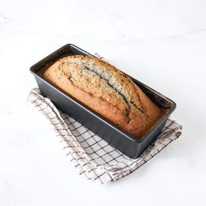 May include: A loaf of bread with a dark brown crust and a light brown interior is in a black loaf pan. The pan is sitting on a white and brown checkered kitchen towel.