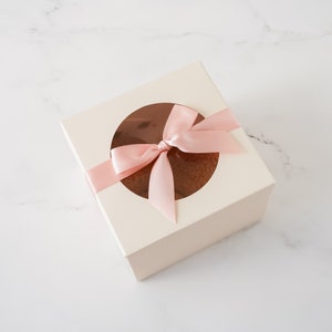 May include: A white gift box with a clear window and a pink satin ribbon tied in a bow. The box is on a white marble surface.