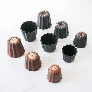 May include: A set of six black and brown fluted cake molds and four baked canelé pastries with a light brown glaze and a purple flower on top.