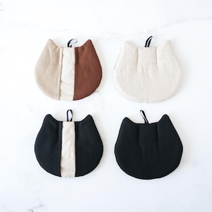 May include: Four cat-shaped pot holders in various colors. One is tan and brown, another is off-white, and the other two are black. Each has a loop for hanging. The pot holders are made of fabric and are designed to protect surfaces from heat.