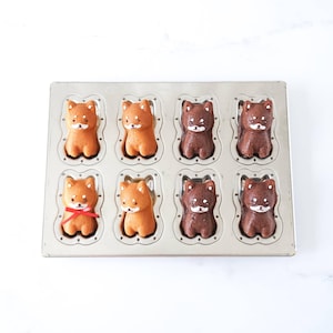 May include: A metal baking pan with eight individual molds shaped like Shiba Inu dogs. The molds are filled with baked goods, some are brown and some are light brown.