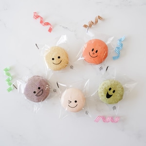 May include: Five colourful macarons with different smiley faces on them are packaged in clear plastic bags with colourful ribbons.