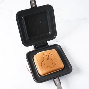 May include: Open black waffle iron with a square waffle featuring a bunny design. The waffle iron is open, revealing the bunny-shaped waffle inside. The iron has a handle and is on a white surface.