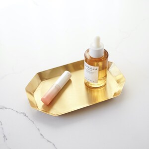 May include: A gold metal tray with a rounded octagonal shape holds a clear glass bottle with a dropper and a pink and white tube. The bottle contains a light yellow liquid and the label reads "Centella Serum".