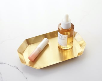 Könnte beinhalten: Ein goldfarbenes Tablett mit abgerundeter achteckiger Form enthält eine klare Glasflasche mit Tropfer und ein rosa-weißes Röhrchen. Die Flasche enthält eine hellgelbe Flüssigkeit und das Etikett lautet "Centella Serum".