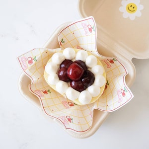 May include: A white and red cherry-themed cake with whipped cream and cherries on top, in a white paper container with a yellow and white checkered pattern.