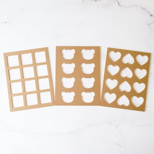May include: Three brown cardboard cookie cutters with different shapes. The first cutter has 16 square cutouts. The second cutter has six bear-shaped cutouts. The third cutter has six heart-shaped cutouts.