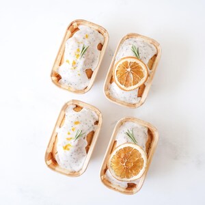Four small rectangular molds with baked goods, orange slices, and herbs on a light background