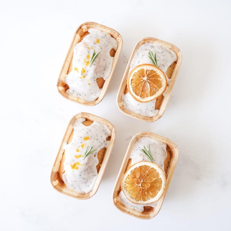 Four small rectangular molds with baked goods, orange slices, and herbs on a light background