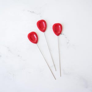 May include: Three red plastic-tipped pins with silver metal shafts. The pins are arranged in a triangular shape on a white background.