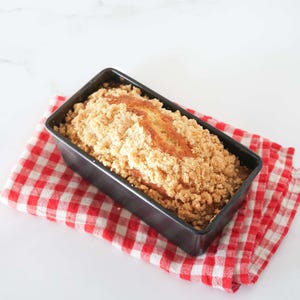 May include: A loaf of bread topped with a crumb topping in a black loaf pan on a red and white checkered kitchen towel.