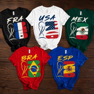 May include: Five short-sleeved t-shirts in black, white, red, green, and blue. Each shirt displays a country's flag design with the country's abbreviation and the number 23. The shirts are arranged on a wooden surface.