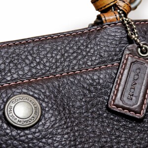 May include: Close-up of a dark brown leather Coach handbag. The bag features a textured surface, pink stitching, and a silver-tone metal button with the Coach logo. A leather tag with the Coach logo hangs from a chain.