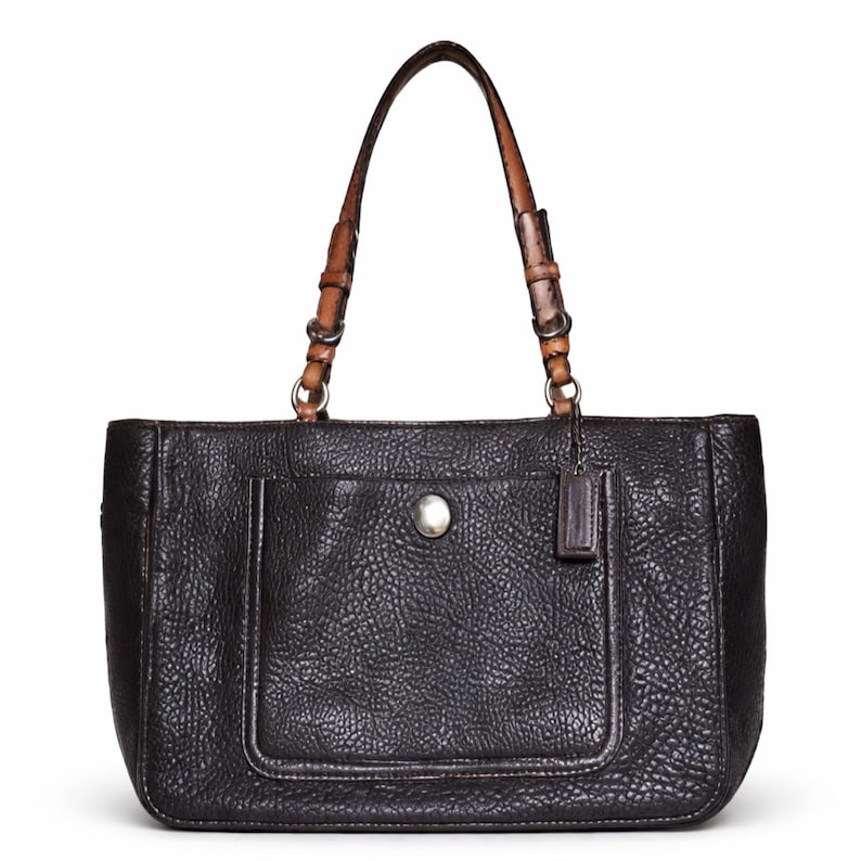 May include: Black leather tote bag with a textured finish. Features a front pocket with a silver button closure and a small leather tag. The handles are brown leather with silver metal accents.