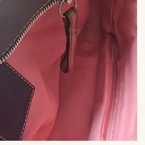 May include: Interior view of a black handbag with a pink lining. A brown leather pull is attached to a silver-colored zipper. White stitching accents the black exterior.