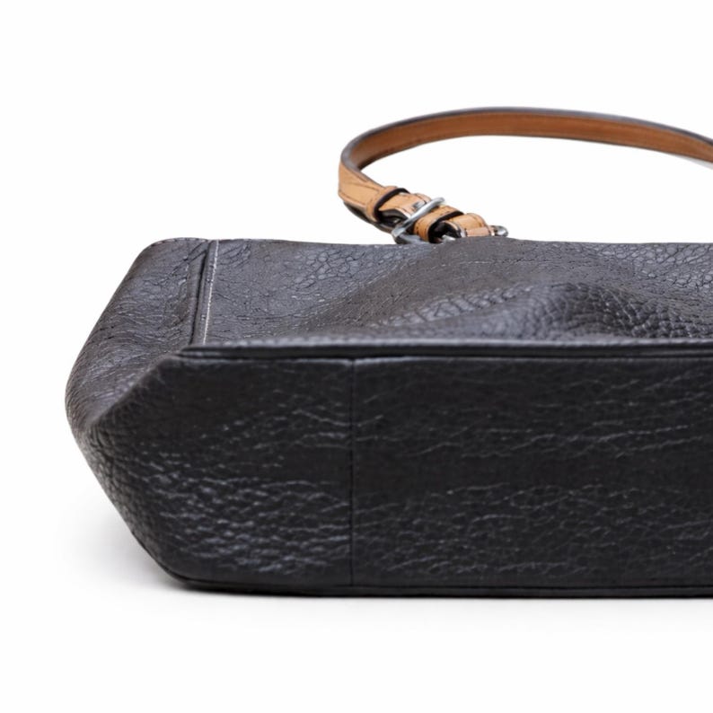 May include: A black leather handbag with a textured surface. The bag has a light brown handle with a silver buckle. The bottom of the bag is rectangular with rounded corners.
