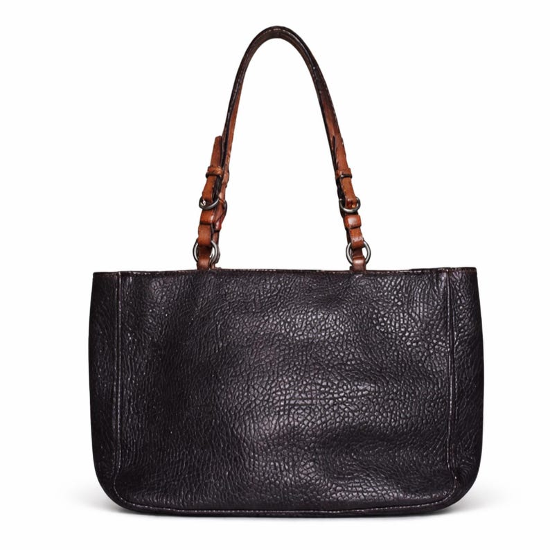 May include: Black leather tote bag with a textured surface. The bag features brown leather handles attached with silver-colored metal rings. The handles are arched and have a lighter brown accent. The bag is rectangular with rounded corners.