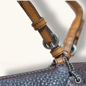 May include: Close-up of a brown leather handbag with a light brown handle and strap. The strap is attached to the bag with silver metal rings and a chain. The bag has pink stitching.