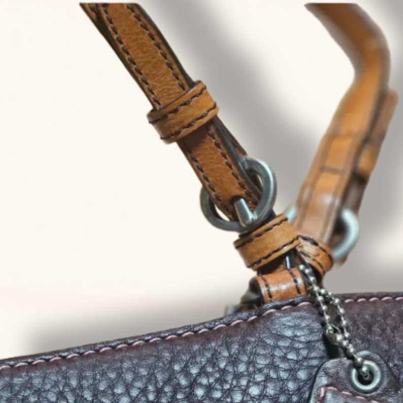 May include: Close-up of a brown leather handbag with a light brown handle and strap. The strap is attached to the bag with silver metal rings and a chain. The bag has pink stitching.