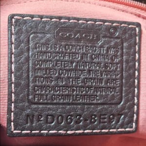 May include: Close-up of a dark brown leather tag with the Coach logo and text. The tag is stitched with pink thread and reads "THIS IS A COACH BAG... HANDCRAFTED IN CHINA... FULL GRAIN LEATHER." The tag also has the code N&deg;D063-8E97.