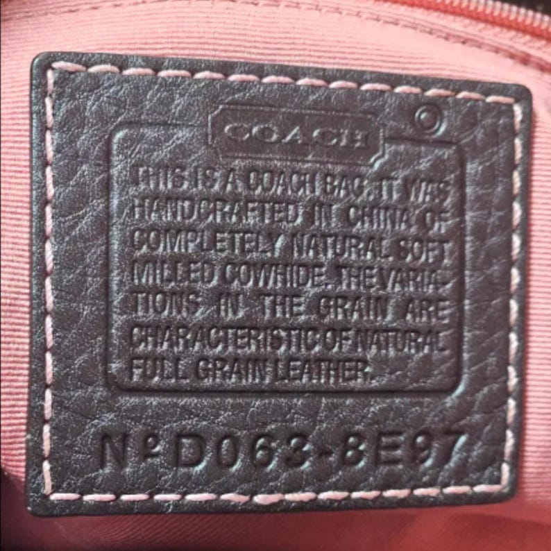 May include: Close-up of a dark brown leather tag with the Coach logo and text. The tag is stitched with pink thread and reads "THIS IS A COACH BAG... HANDCRAFTED IN CHINA... FULL GRAIN LEATHER." The tag also has the code N&deg;D063-8E97.