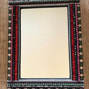 May include: Rectangular mirror with a mosaic frame. The frame features a pattern of red, green, and silver accents. The top and bottom edges have silver-colored embellishments. The mirror is a decorative item.