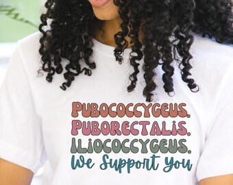 May include: White t-shirt with a colorful graphic that reads "PUBOCOCCYGEUS. PUBORECTALIS. ILIOCOCCYGEUS. We Support you".