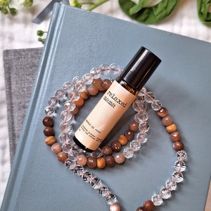May include: A blue notebook with a clear crystal and brown stone mala necklace and a small brown glass bottle with a white label that reads "relaxed aroma" on it.