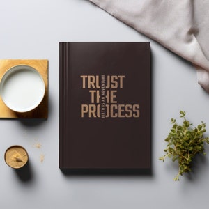 May include: A dark brown journal with the text "TRUST THE PROCESS" in gold lettering. The text "BIRTH IS AN ADVENTURE" is in smaller gold lettering below the main text.