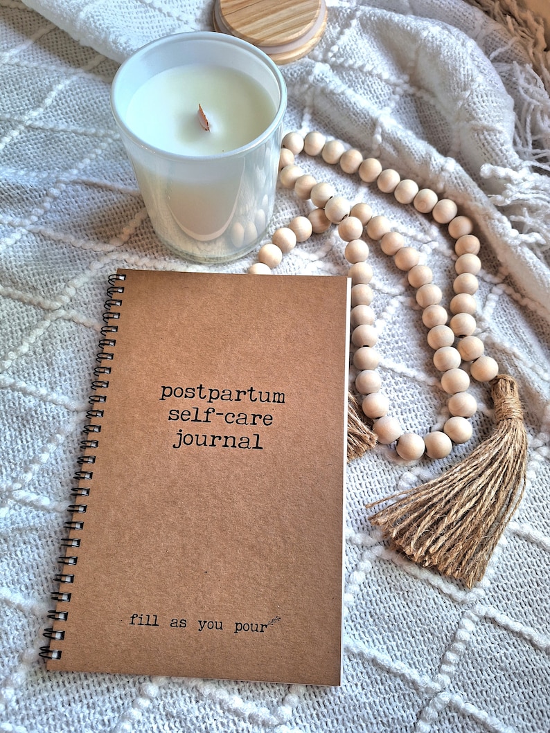 May include: A brown spiral bound journal with the text "postpartum self-care journal" and "fill as you pour" on the cover. The journal is on a white blanket with a white candle and a wooden bead garland with a tassel.