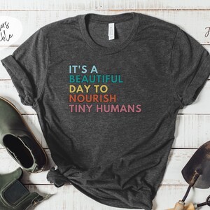 Puede incluir: Camiseta gris oscuro con el texto "IT'S A BEAUTIFUL DAY TO NOURISH TINY HUMANS" en turquesa, amarillo, naranja y rosa. La camiseta se muestra con herramientas de jardiner&iacute;a y botas verdes.