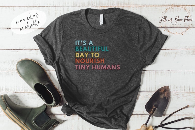 Puede incluir: Camiseta gris oscuro con el texto "IT'S A BEAUTIFUL DAY TO NOURISH TINY HUMANS" en turquesa, amarillo, naranja y rosa. La camiseta se muestra con herramientas de jardiner&iacute;a y botas verdes.