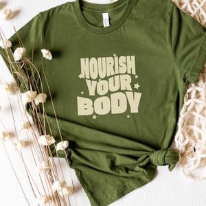 Puede incluir: Camiseta verde con el texto "Nourish Your Body" en una fuente retro con estrellas alrededor de las palabras.